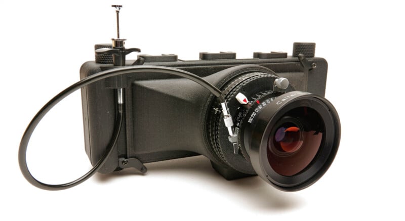 A black, box-shaped camera with a large lens and a cable release attached, placed on a white background.