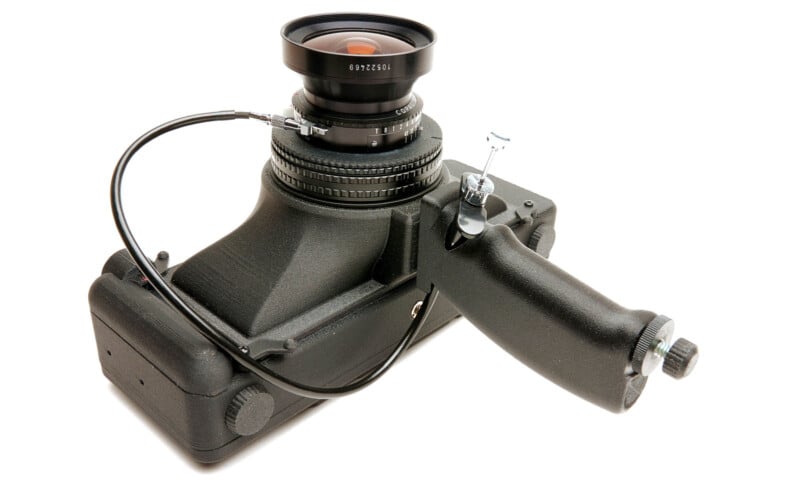 A black, handheld medium format film camera with a large lens, trigger grip, and cable release attachment, set against a white background.
