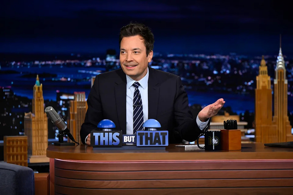 Jimmy Fallon in a suit smiles while seated behind a desk with buttons labeled "This" and "That" with a cityscape background behind him