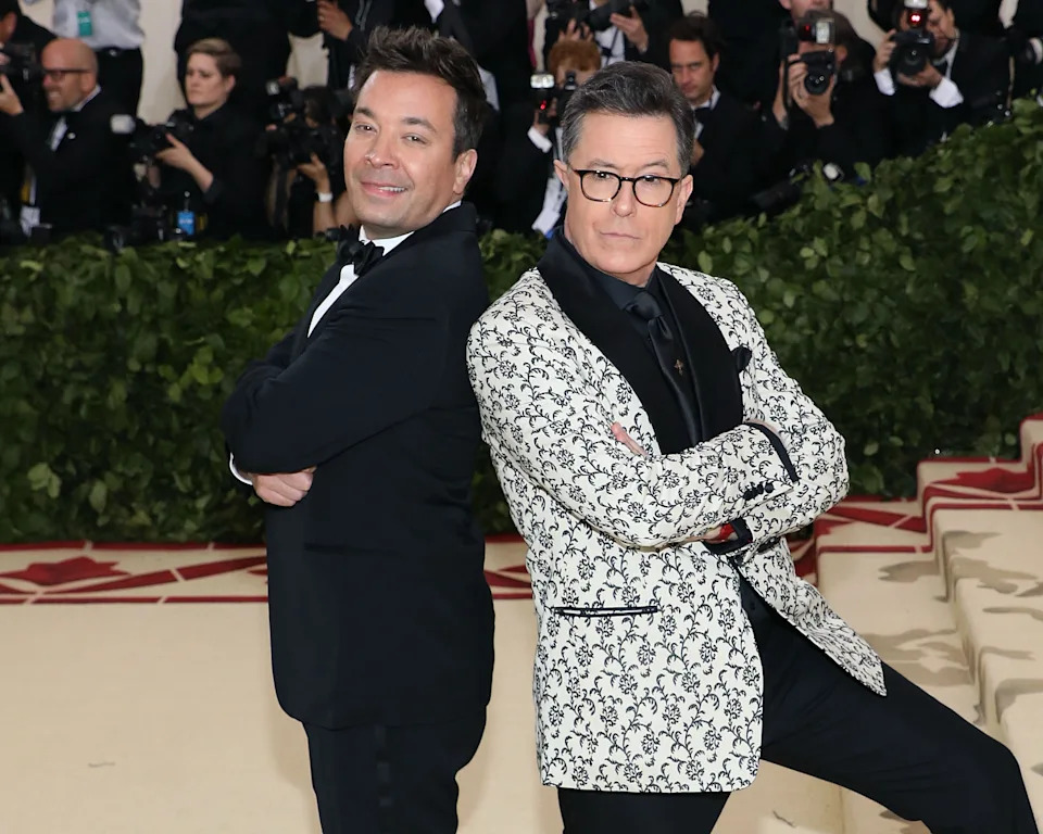 Jimmy Fallon and Stephen Colbert pose with their backs against each other on a red carpet
