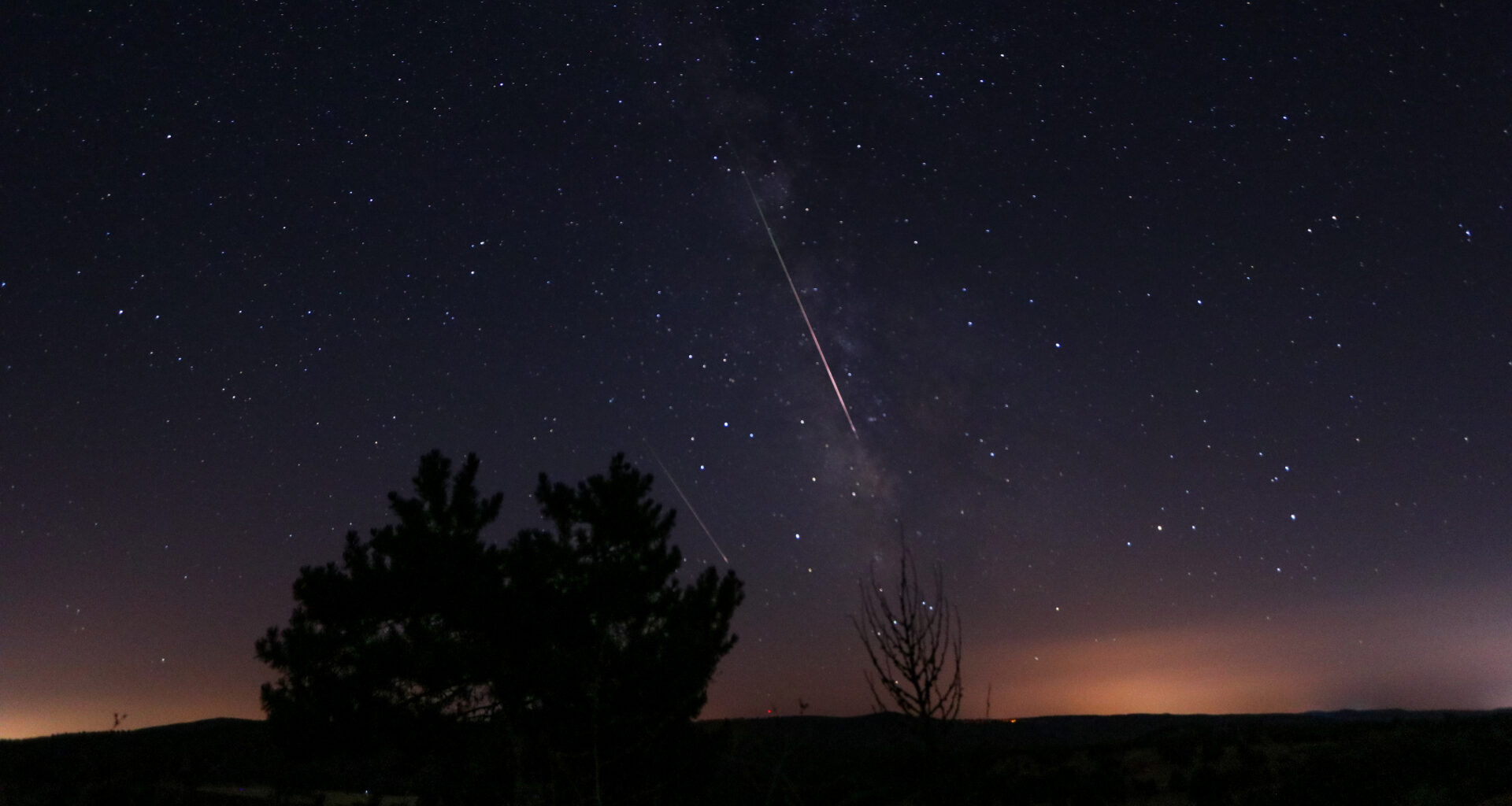 Two meteors are pictured racing through a starry night sky parallel to the Milky Way. Light pollution is visible above a silhouetted horizon and trees.