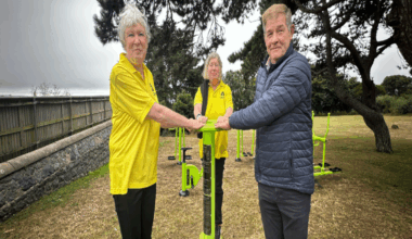 Two woman wearing yellow bowel cancer guernsey shirts and a man wearing a blue puffer jacket holding onto a lime green piece of exercise equipment. They are outside and standing in front of a group of trees.