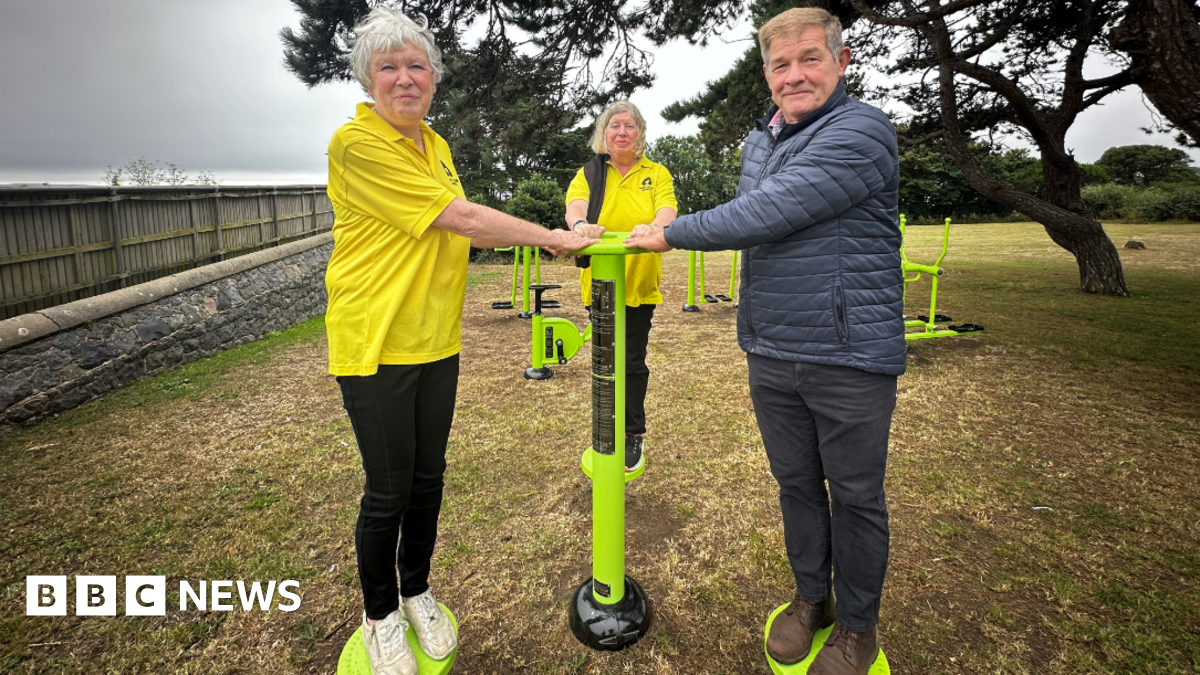 Two woman wearing yellow bowel cancer guernsey shirts and a man wearing a blue puffer jacket holding onto a lime green piece of exercise equipment. They are outside and standing in front of a group of trees.