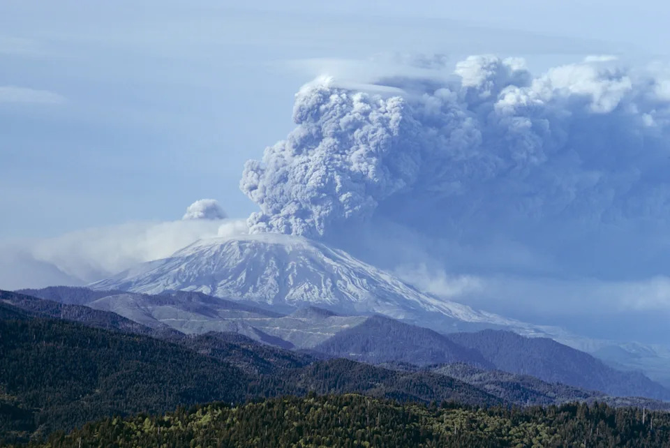 Volcano erupting with large plumes of ash and smoke billowing into the sky, surrounded by forested mountains in the foreground