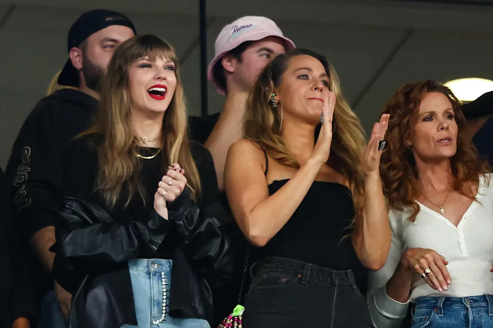Three women at an event, with one in a leather jacket and another in a strapless top, clapping and smiling. Crowd in the background