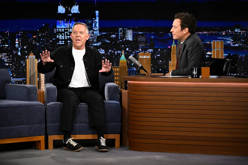 Greg Gutfeld in a casual outfit and sneakers talks animatedly while seated on a talk show set, facing Jimmy Fallon behind a desk