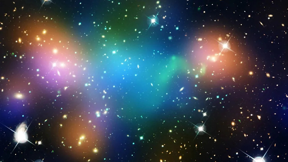  Orange, blue, purple, and green bursts of light are shown over the glow of stars in a deep space image. 