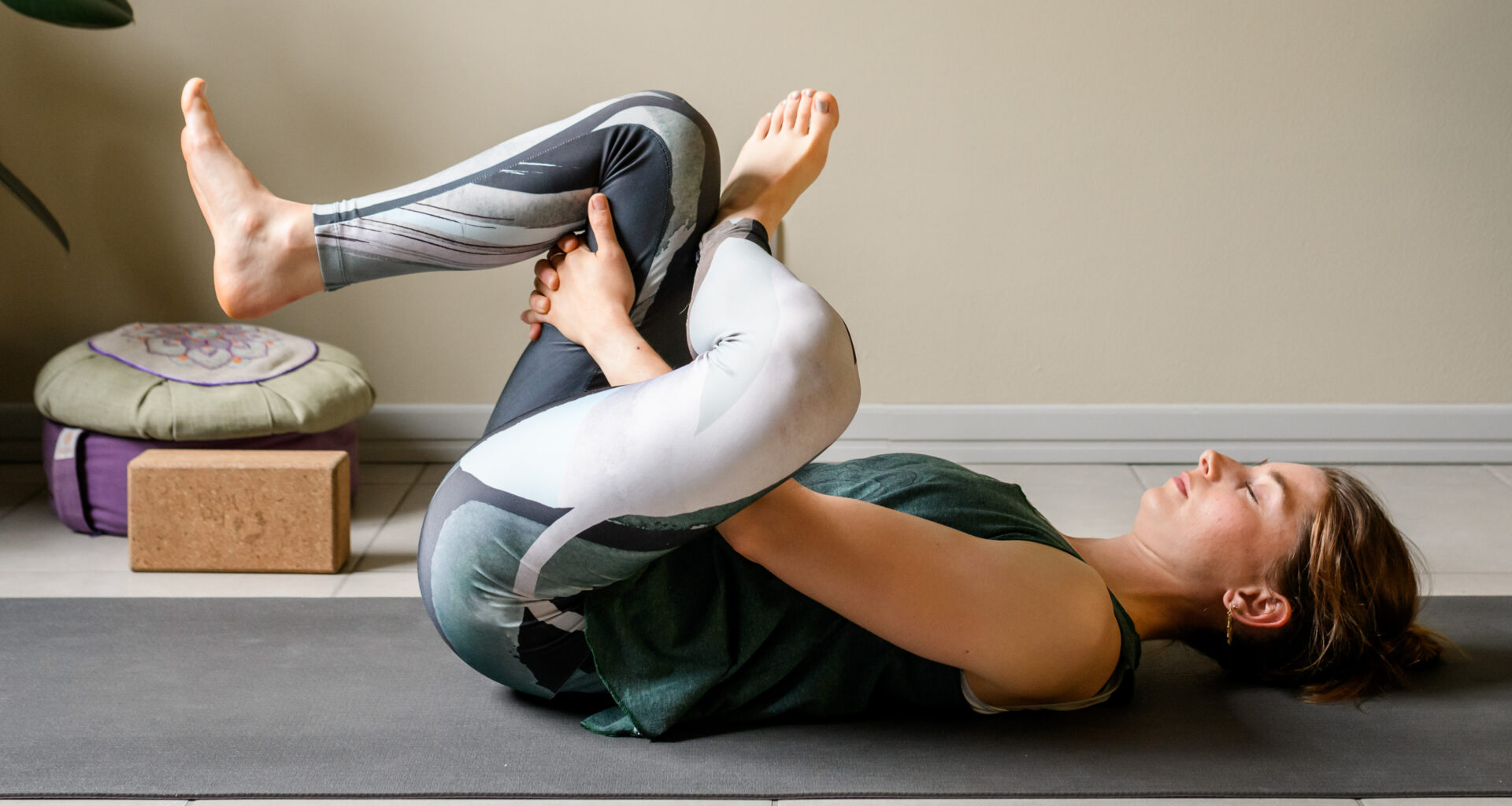 A woman practices a figure four yoga stretch at home, reclining on her back. Her eyes are closed and we see a yoga block and cushion behind her.