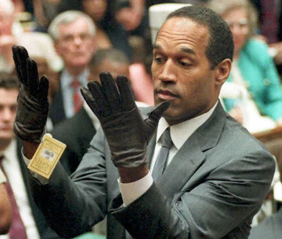 A man in a suit tries on leather gloves during a public event, surrounded by onlookers in formal attire