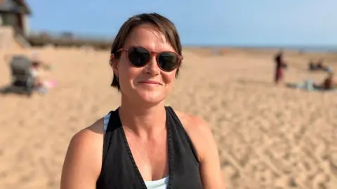 A woman sitting on the beach and smiling at the camera. She is wearing a black halter neck top and sunglasses. A sandy beach is in the background.