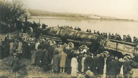 MNH Sepia photograph of crowds of people look on as the dead whale is transported. Derbyhaven can be seen in the background.