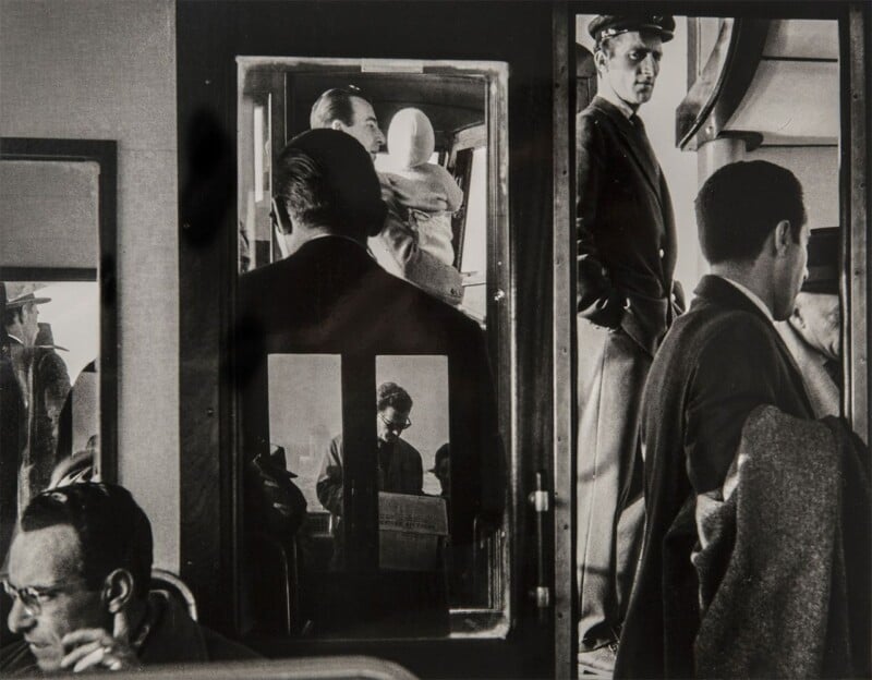A black and white photo shows several men in suits and hats standing and sitting inside and around a train or bus. Reflections in glass doors create layered images of the figures.