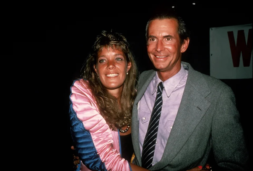 Two people smiling and posing together at an event. The woman wears a shiny jacket, and the man is in a suit