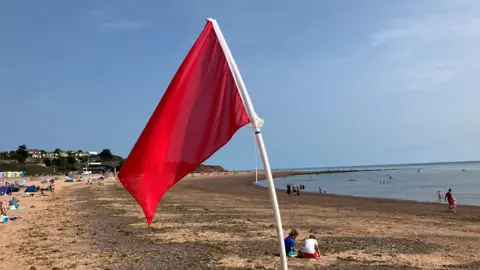 BBC A red flag is blowing in the wind on a beach. Beachgoers are in the distance. Swimmers are in the water. The sky is blue.