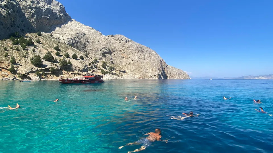 A turquoise sea below a light-coloured cliff face. In the sea, there are several people swimming, spread out across the image. In the background, there is a red, large boat. 