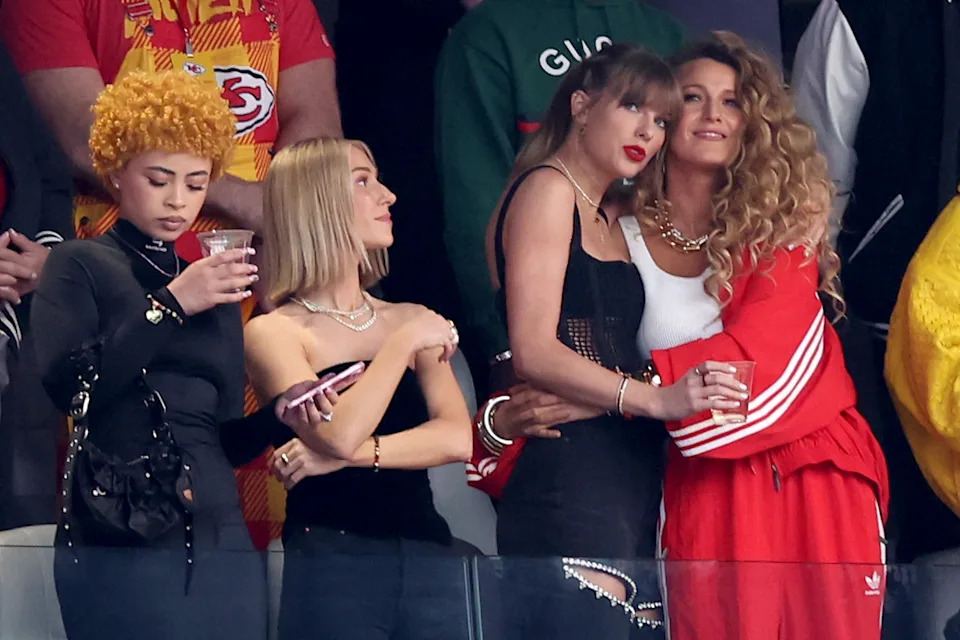 Four women at an event: one with yellow curls and drink, one in black sleeveless, another in a black dress, and one with long curls in a red tracksuit