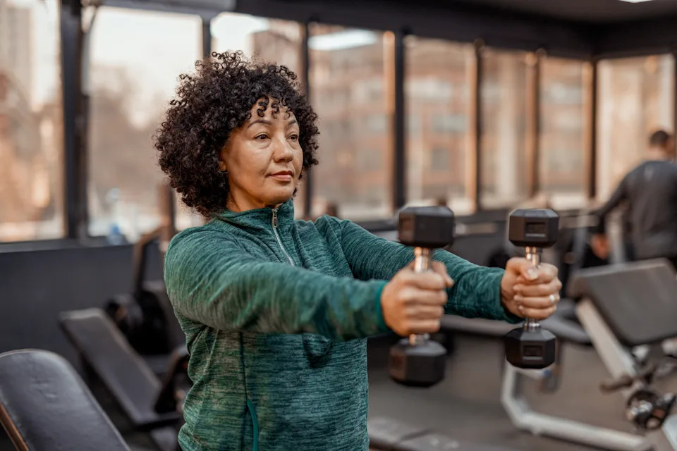 A Middle-aged, Plus-size Woman Performing an Upper Body Workout With Dumbbells in the Gym. She is Seen Holding a Dumbbell in Each Hand, Focusing on Her Form and Breathing. Her Face Shows a Mix of Effort and Satisfaction as She Continues Her Workout Routine. The Background Features Various Gym Equipment, Reinforcing the Environment of Fitness and Well-being. The Image Highlights That Fitness is Inclusive and for Everyone, Encouraging Individuals of All Ages and Body Types to Take Care of Their Health.