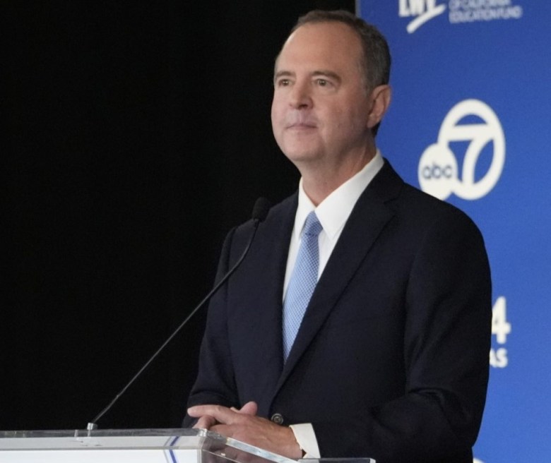 Man in a suit speaking into a microphone with a blue and black background.