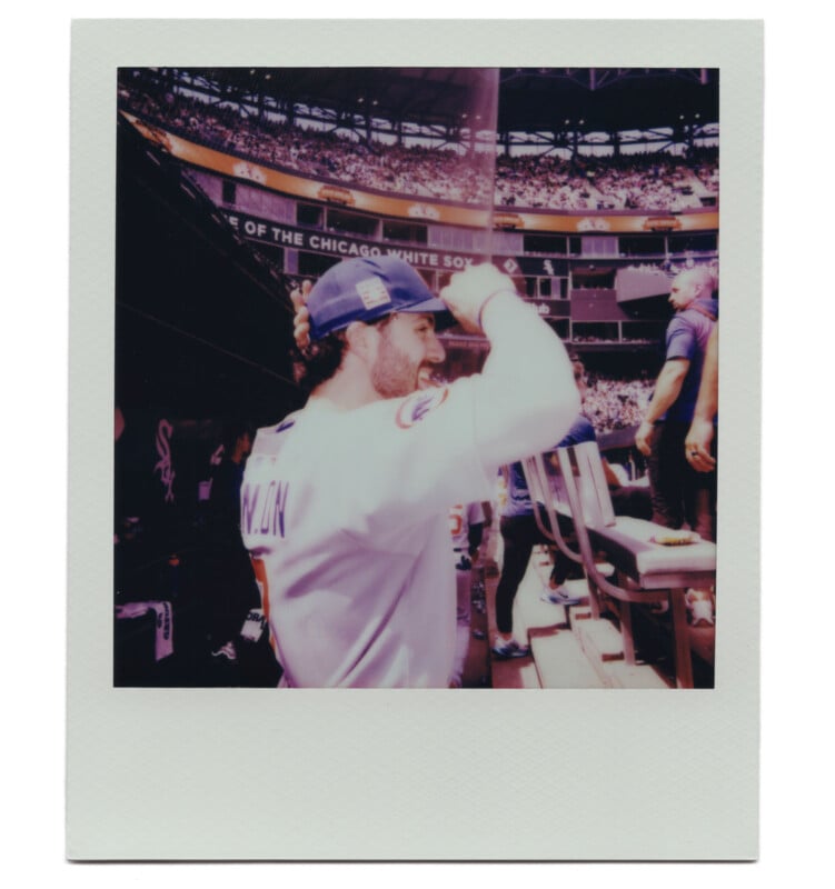 A man in a baseball jersey salutes and smiles while standing near the stands at a baseball stadium, with fans and the field visible in the background.