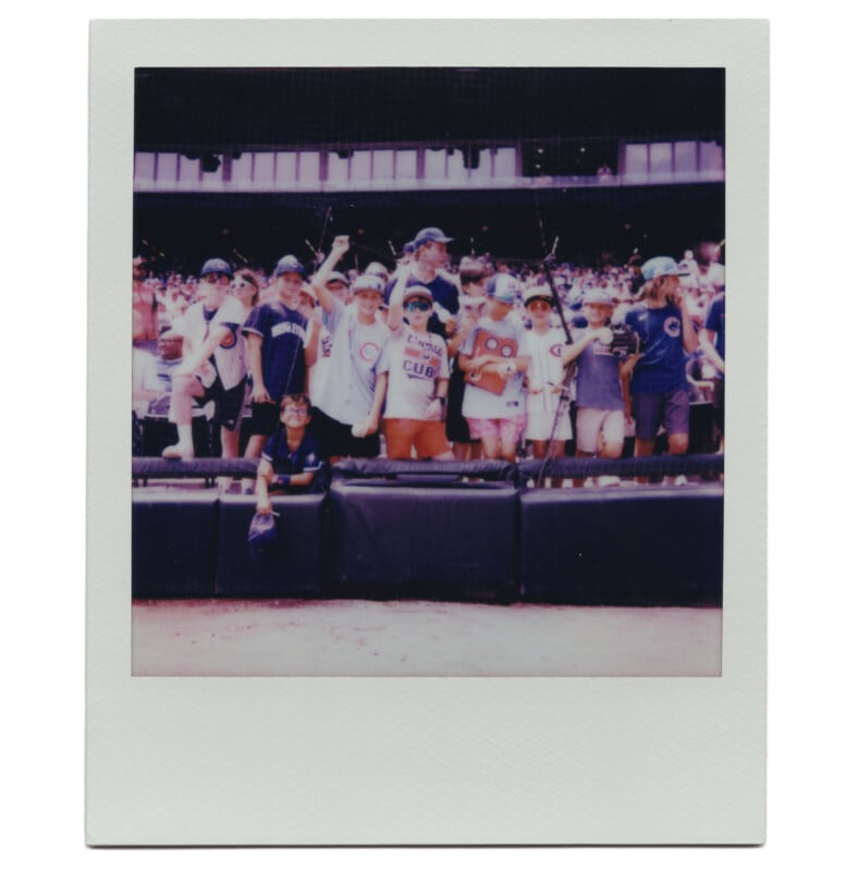 A group of children wearing baseball caps and Cubs shirts pose and smile together by the railing at a baseball stadium, with a crowd of fans visible in the background.
