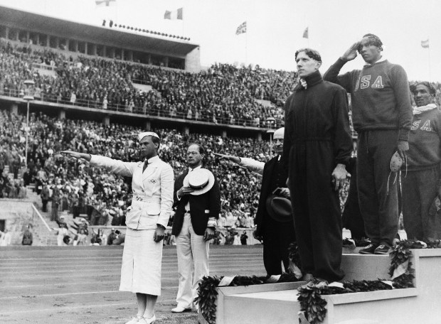 In this Aug. 3, 1936, file photo, gold medalist Jesse Owens, second right, salutes during the playing of the national anthem during the medal ceremony of the 100-meter final in Berlin. Silver medalist Tinus Osendarp, third from right, of, Holland, and bronze medalist Ralph Metcalf, right, listen, along with a matron who holds a Nazi salute. The 1936 Games in Berlin, awarded about two years before Adolf Hitler became dictator, went ahead under Nazism. American track great Jesse Owens went on to win four gold medals, but he was only supposed to compete in three events, the 100 meters, 200 meters and long jump. (AP)