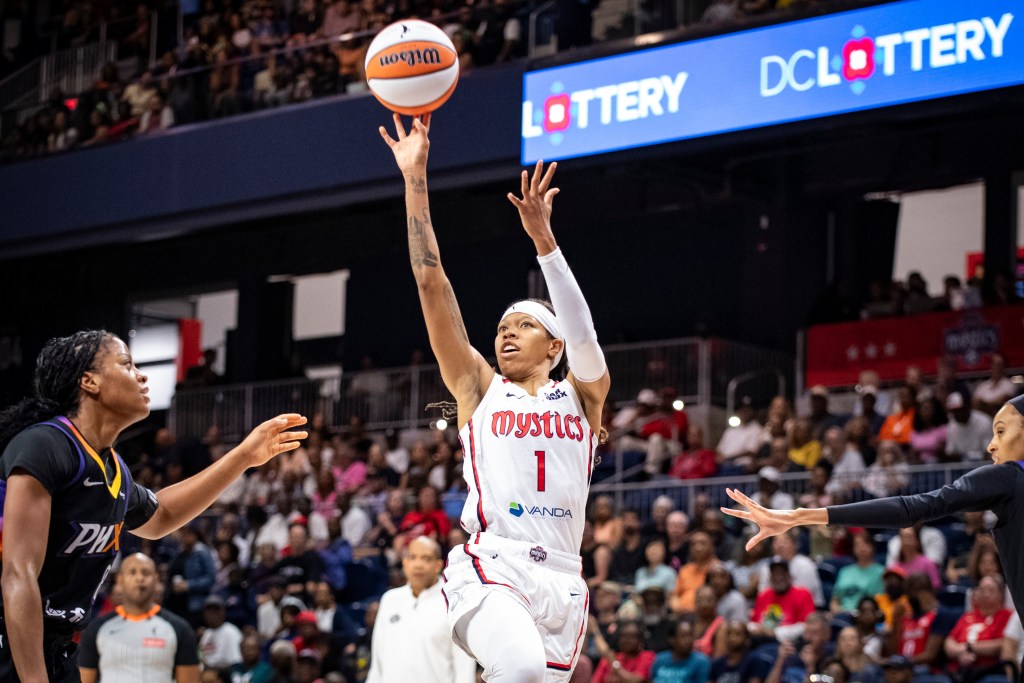 Washington Mystics guard Sug Sutton shoots a right-handed floater off her left leg. Two Phoenix Mercury defenders reach their arms toward her, but neither is close enough to contest the shot. Mystics head coach Sydney Johnson is visible in the background behind Sutton.