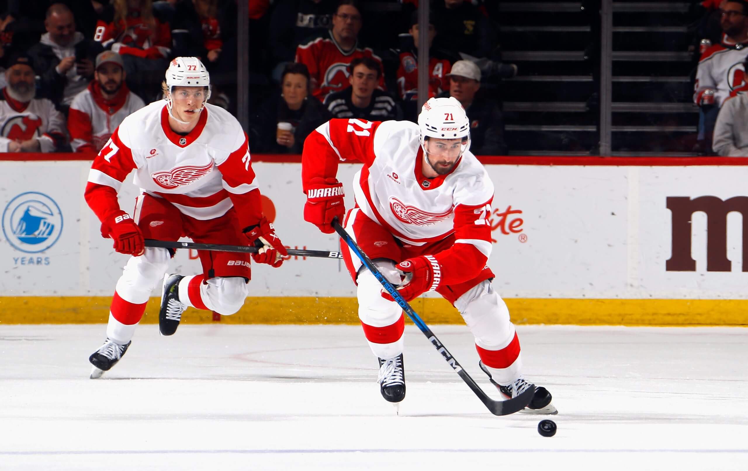 The Red Wings' Dylan Larkin chases after the puck with teammate Simon Edvinsson in the background of the frame.