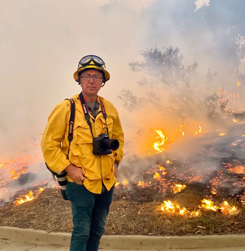 A person wearing a yellow fire-resistant jacket, helmet, and glasses stands with cameras around their neck in front of a smoky, active wildfire with flames and burning vegetation.