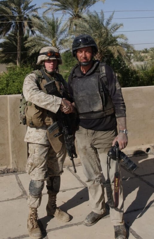 A soldier in camouflage uniform and helmet shakes hands with a man in black protective gear holding a camera. They stand outdoors near a wall, with palm trees and greenery in the background.