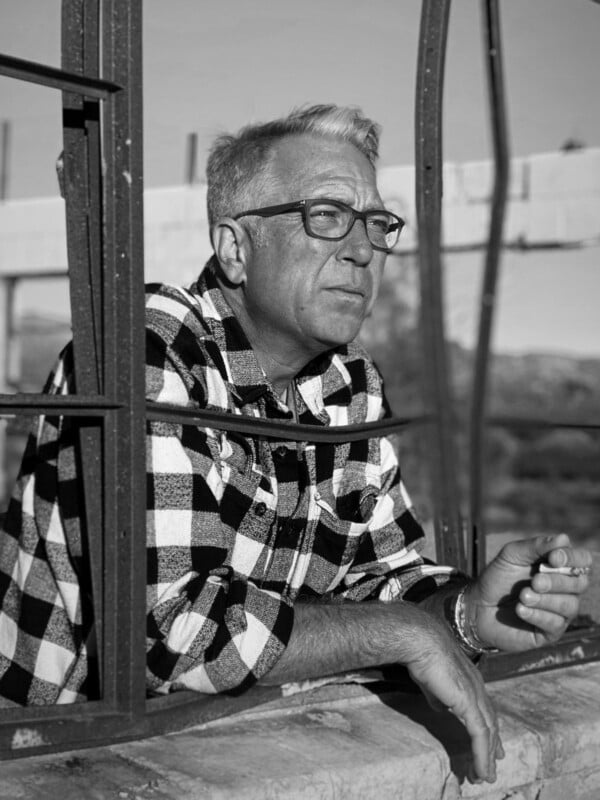 A man with short gray hair and glasses, wearing a plaid shirt, leans on a window frame holding a cigarette. He looks thoughtful, gazing into the distance. The photo is black and white.