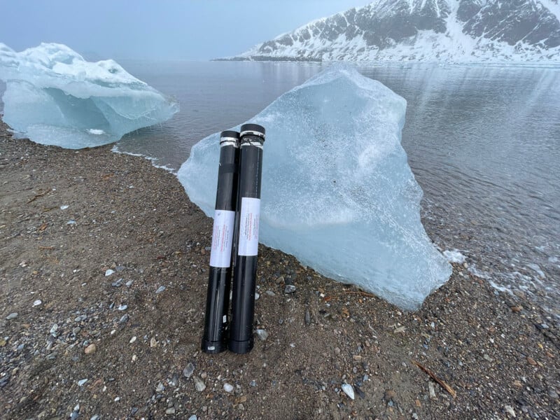Two black cylindrical scientific instruments stand upright on a pebbled shore with ice chunks nearby, next to calm water and snowy mountains in the background.