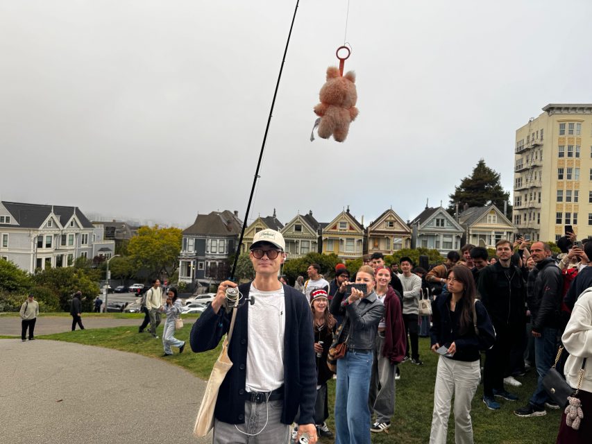 A person holds a fishing rod with a stuffed animal hanging from the line in front of a crowd at a park, with Victorian houses in the background.