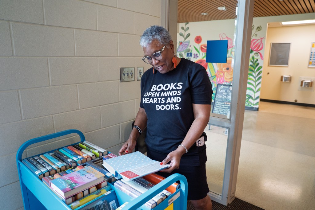 A Black woman with short salt-and-pepper hair, wearing a shirt that reads, "Books open minds, hearts and doors," smiles and looks through a book on a book cart.