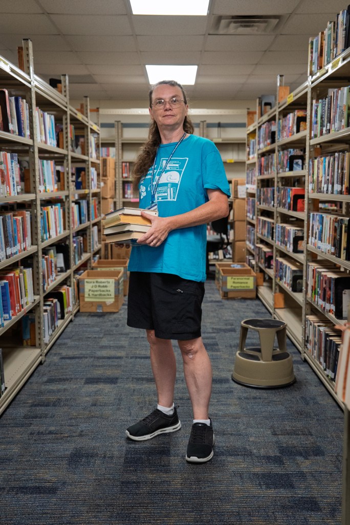 A person, wearing a bright, light blue T-shirt hold a stack of books and poses for a portrait in a library aisle.
