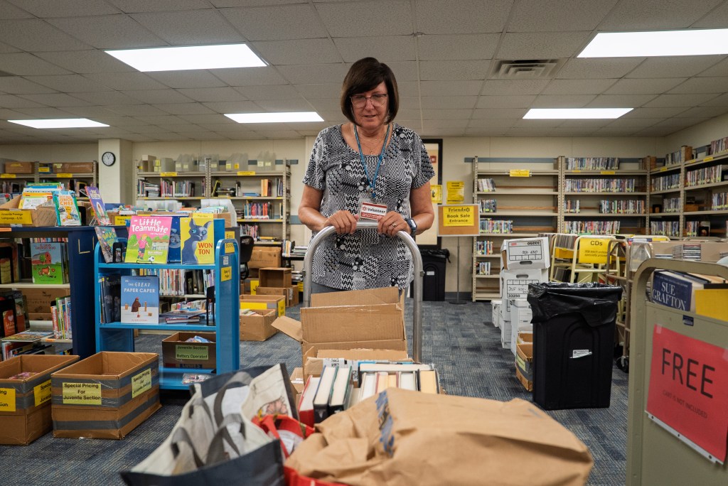 A white woman with a brown bob haircut, wearing a patterned shirt moves a metal caddy filled with books through a library.