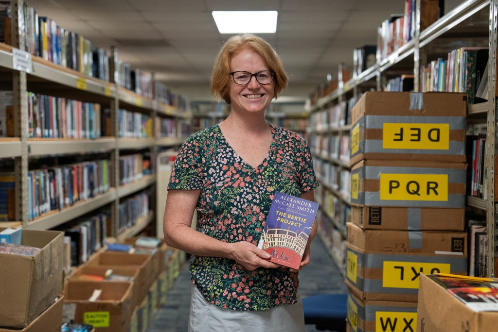 A white woman, wearing a flower-patterned shirt and glasses, holds a book and smiles as she poses for a photo in a library book aisle.
