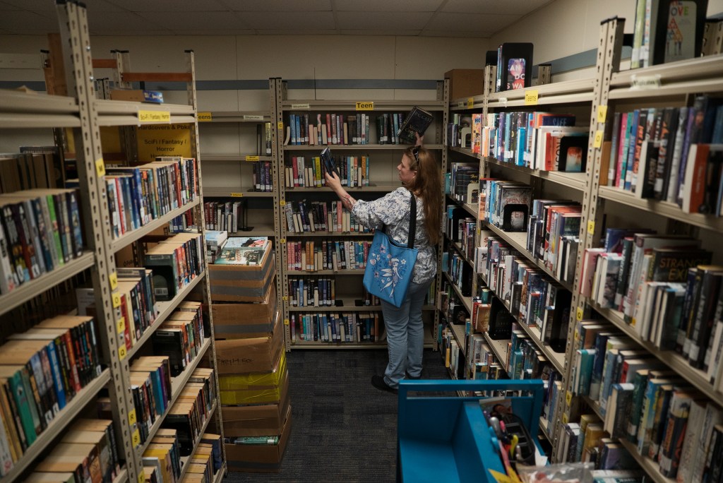 A white woman with long brown hair in casual clothing browses through a library bookshelf.