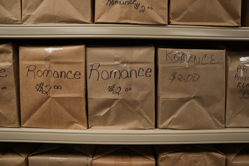 A bookshelf holds boxes or brown wrapping paper with "Romance" and "$2.00" written on the sides of each of them.