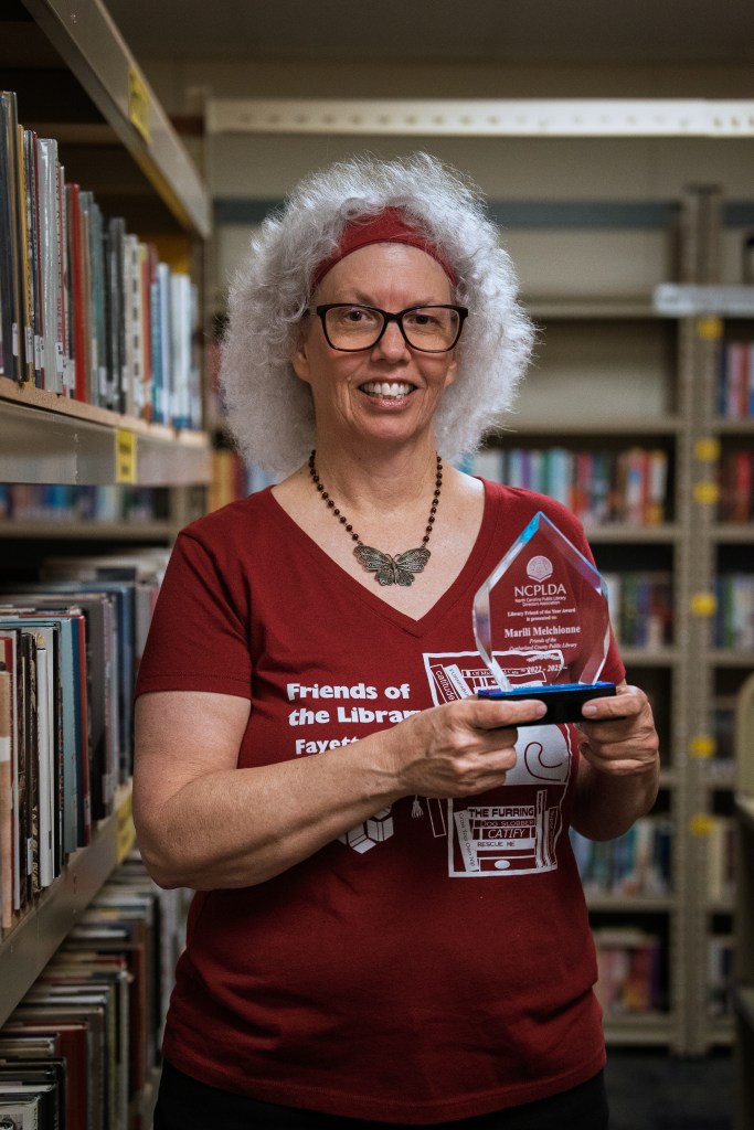 A woman in a maroon shirt holds an award in the library