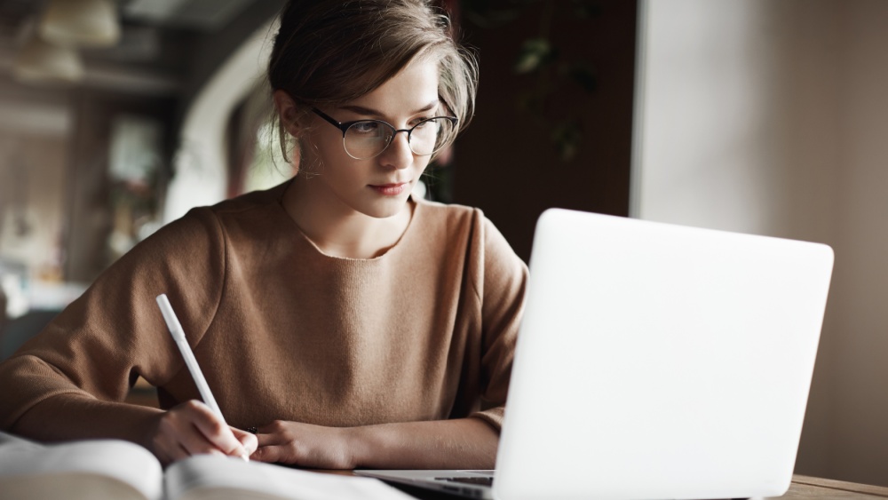 A person is sitting in front of her open PC and writing on a paper.
