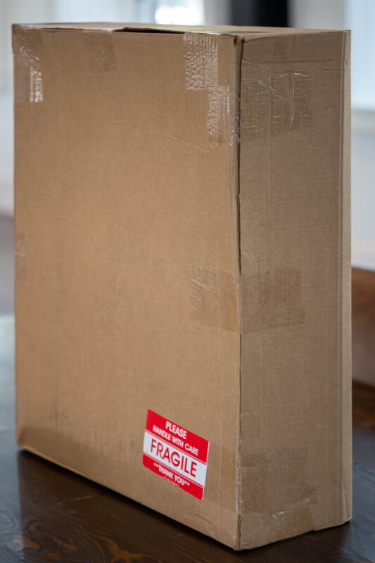 A large, sealed cardboard box with a red and white label reading "Please handle with care. FRAGILE. Thank you." sits on a wooden surface indoors.