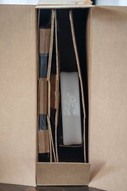 Open cardboard box with protective cardboard dividers and a piece of white foam wrapped in plastic inside, viewed from above.