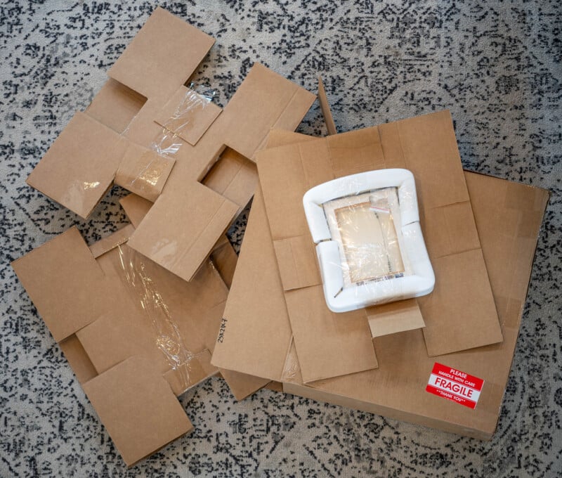 Cardboard pieces and packaging foam lie on a patterned carpet, with a boxed item labeled "fragile" resting on top of larger cardboard sheets.