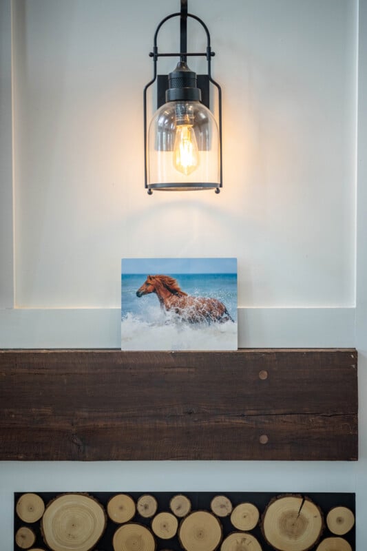 A lit wall sconce hangs above a small painting of a brown horse running through ocean waves, which is displayed on a white wall with rustic wooden accents and a log slice decoration.