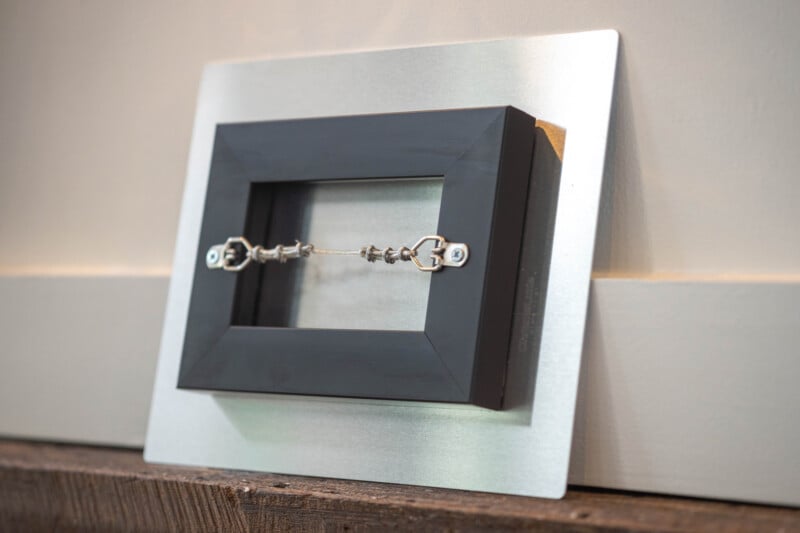 A small, black picture frame is mounted backward on a metallic plate, showing its wire hanging hardware and metal hooks, resting against a beige wall and wooden surface.