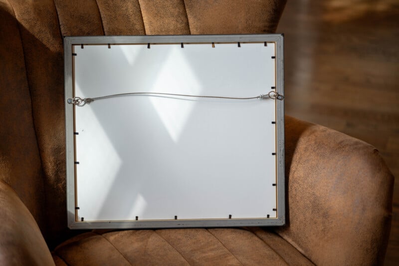 A square picture frame with a hanging wire is resting face down on a brown, textured armchair. Sunlight creates an X-shaped shadow on the white backing of the frame.
