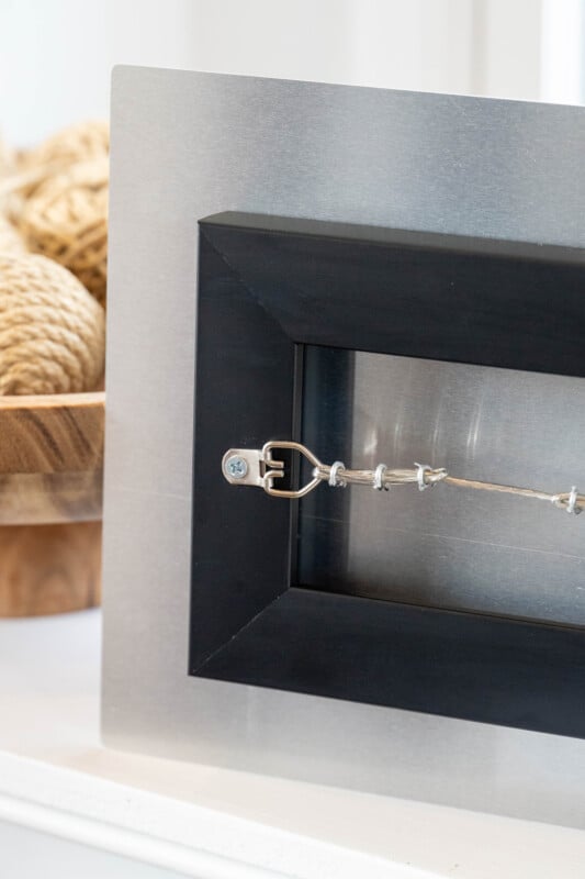 Close-up of the back of a black picture frame with a hanging wire, displayed upright. In the background, there is a wooden bowl filled with twine.