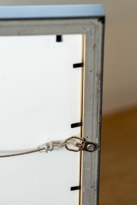 Close-up of the back corner of a picture frame, showing a metal hanging wire attached to a metal loop secured by a screw. The frame's edge and some clips holding the backing are visible.