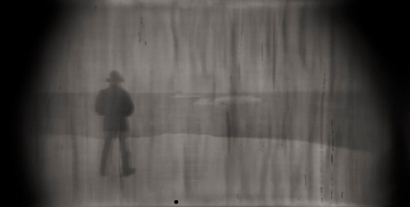 A blurry, black-and-white photo shows a person wearing a hat standing near water, seen through streaked, foggy glass or film, creating a mysterious and vintage atmosphere.