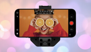 A smartphone on a mount displays a woman holding two orange halves over her eyes and smiling, with an orange background, while the phone screen shows camera controls.